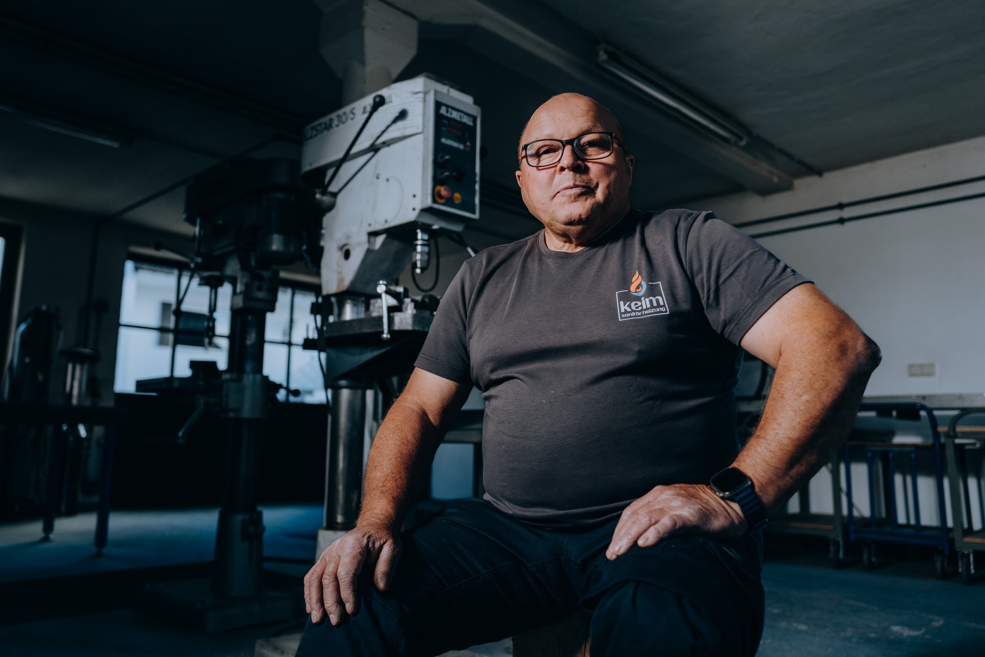 Senior aus dem keim-Team als Werkstattporträt vor der Ständerbohrmaschine.