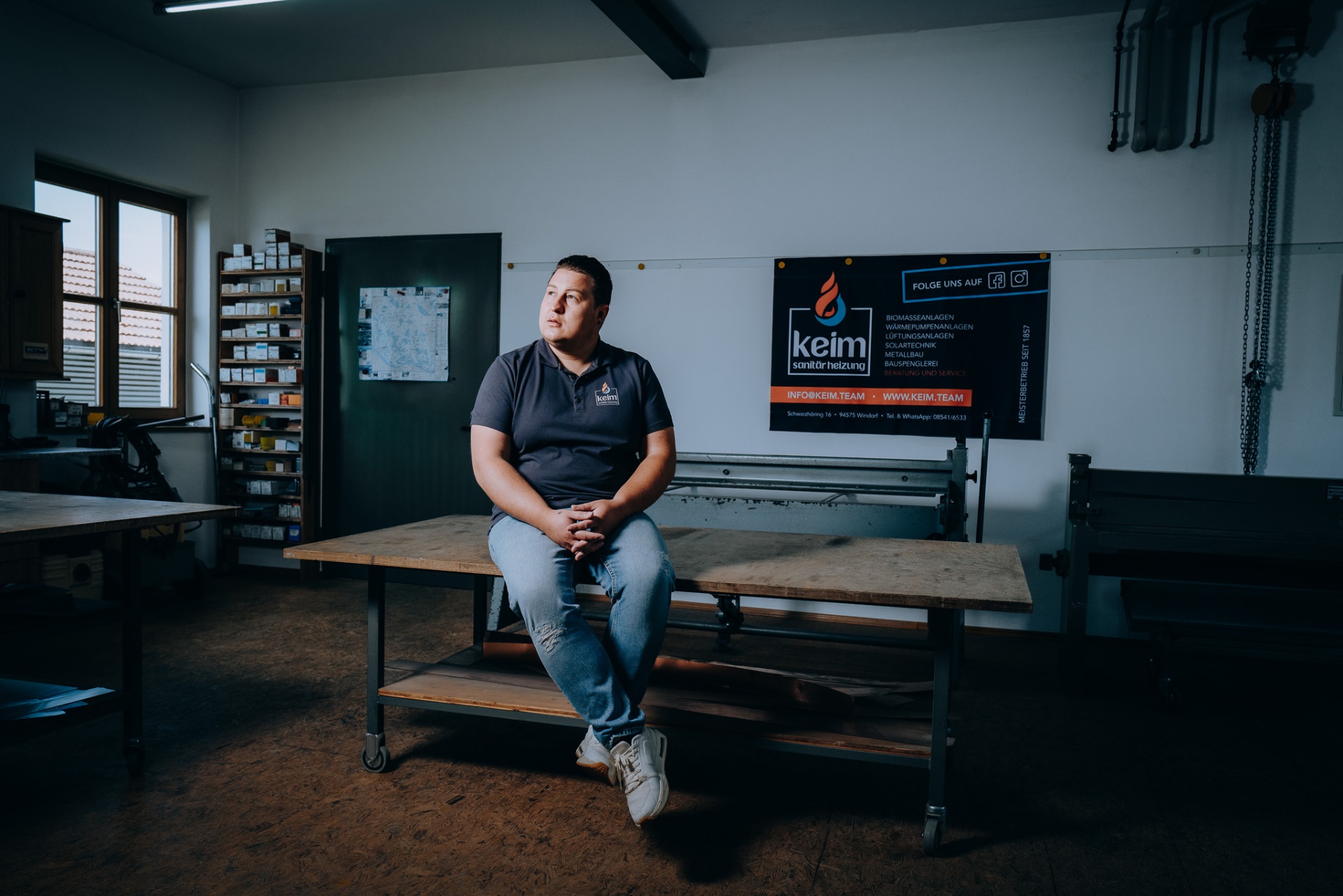 Christoph Keim sitzt in der Spenglerei-Werkstatt und blickt nach links, im Hintergrund das keim-Leistungsbanner.