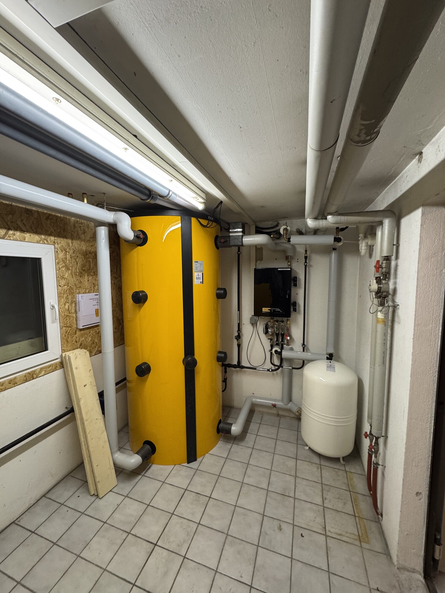 Heizraum mit gelbem Pufferspeicher, Frischwasserstation, Ausdehnungsgefäß und neu geführten Rohrleitungen