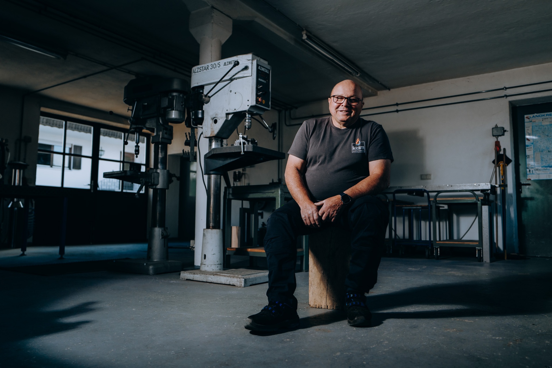Senior-Chef von keim lächelt in der weiten Spenglerei-Werkstatt neben der Bohrmaschine.