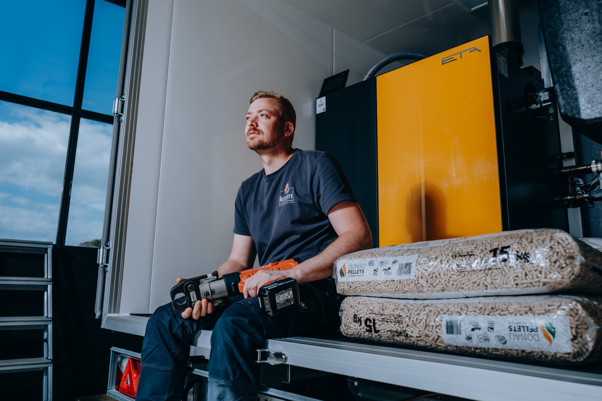 keim-Monteur mit seitlichem Blick im Heizmobil, Presswerkzeug in der Hand und Pelletsäcke im Vordergrund.