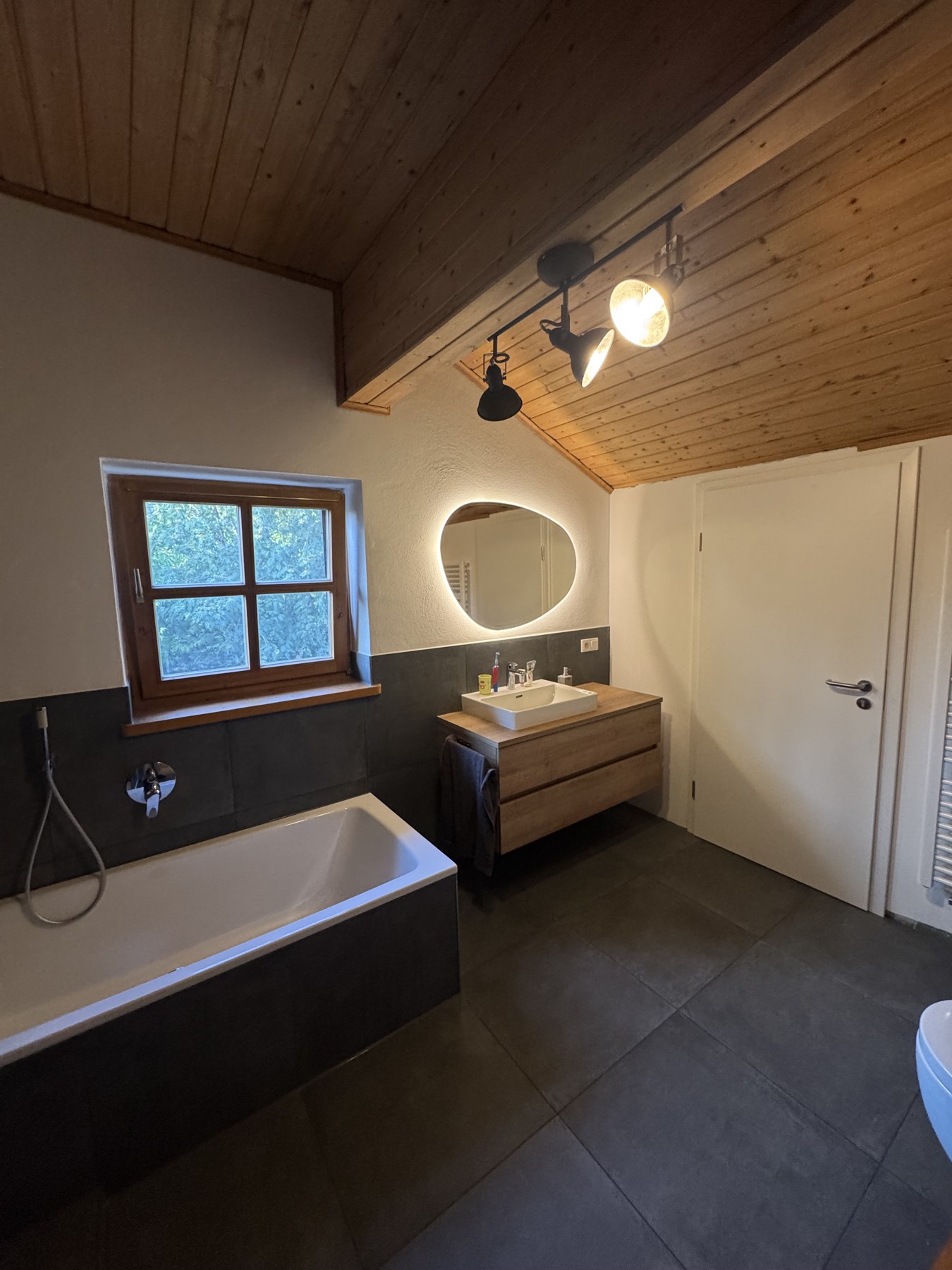 Fertiges Dachbad mit Badewanne, dunklen Großformatfliesen und Holzdecke