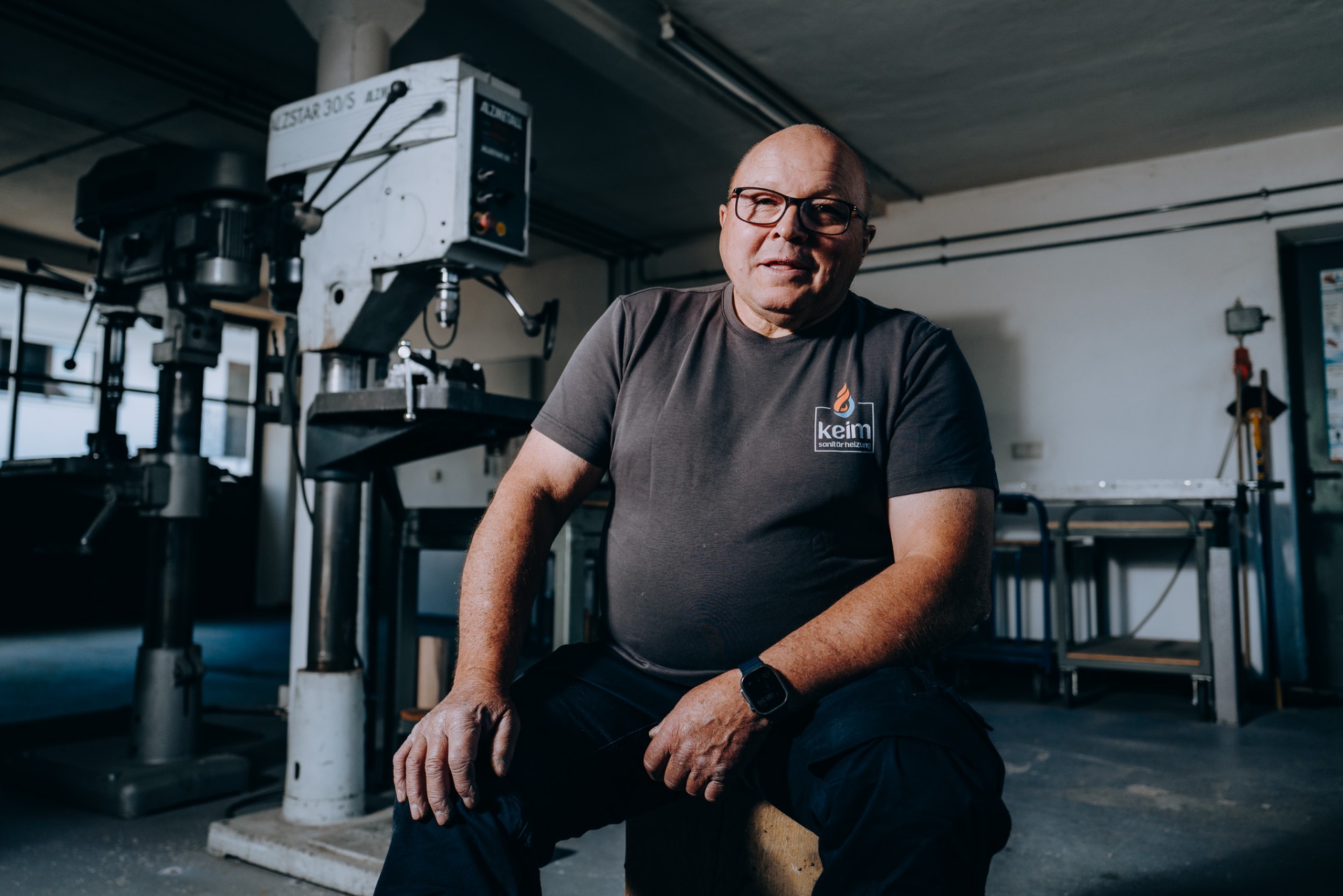 Senior-Chef von keim mit Keim-Logo-Shirt vor der Alzstar-Ständerbohrmaschine, Untersicht.